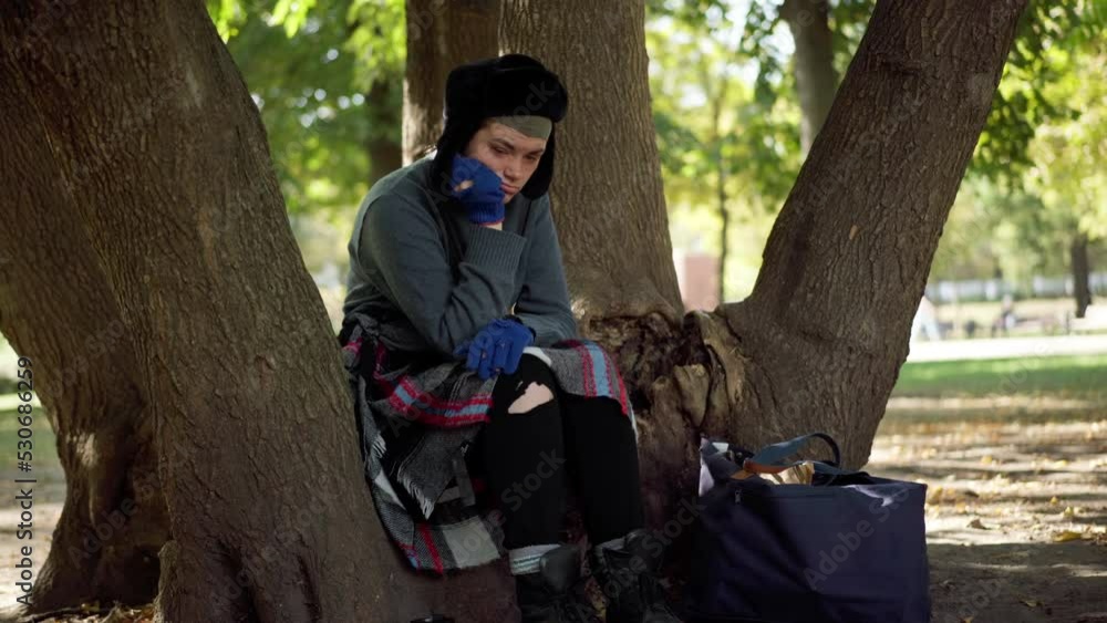 Frustrated homeless woman sitting on tree in park thinking gesturing. Portrait of devastated Caucasian houseless person in urban city park. Social issues concept Frustrated homeless woman sitting on tree in park thinking gesturing. Portrait of devastated Caucasian houseless person in urban city park. Social issues concept