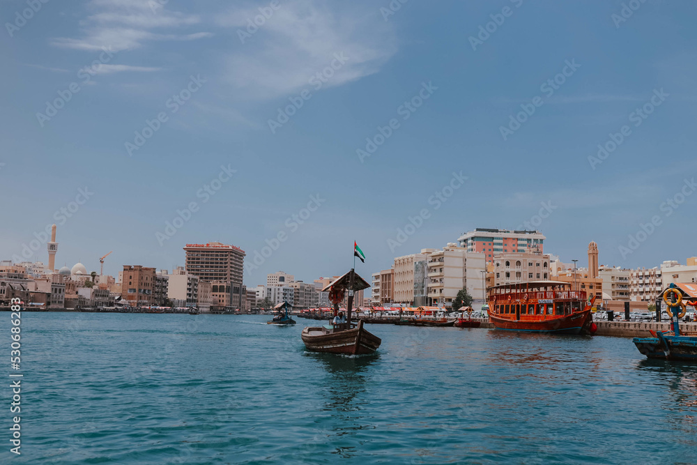 Obraz premium Dubai, UAE - April 15, 2021: Cityscape view of Dubai Creek channel with old wooden traditional boats cruising and piers. Sunny summer day. Famous tourist destination in UAE, United Arab Emirates. Abra