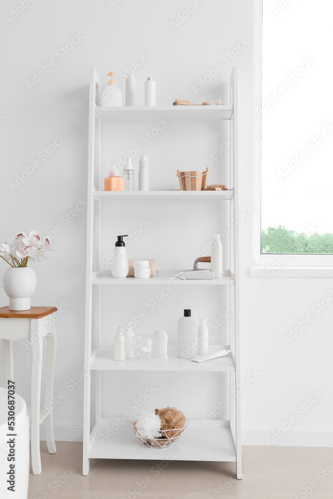 Shelf unit with different bath accessories and vase with flowers on table near white wall