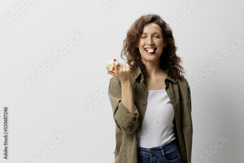 Healthy Lifestyle. Funny smiling curly beautiful woman in casual khaki green shirt eat apple holds piece in mouth posing isolated on white background. Natural Eco-friendly products concept. Copy space