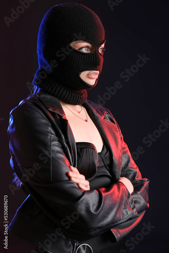 Young woman in balaclava and leather jacket standing with crossed hands on black background