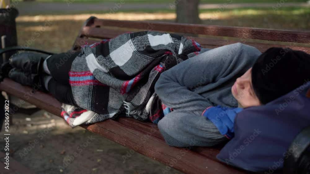 Young homeless woman sleeping lying on bench in urban city park. Wide shot of Caucasian sad helpless poor person outdoors on sunny summer day Young homeless woman sleeping lying on bench in urban city park. Wide shot of Caucasian sad helpless poor person outdoors on sunny summer day