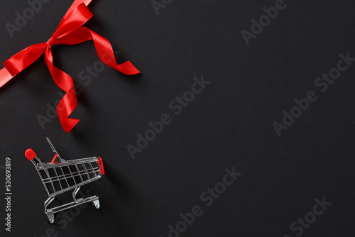 Shopping cart on background of black gift card with red ribbon bow. Black Friday sale banner template.