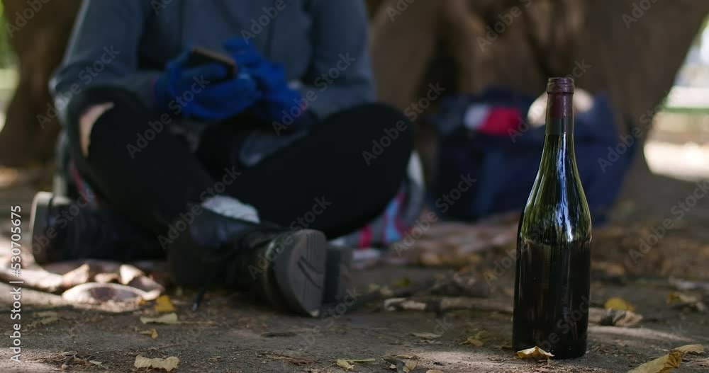 Wine bottle outdoors in city park with unrecognizable blurred homeless woman using phone at background in slow motion. Caucasian young alcohol addicted person living on street. Cinema 4k ProRes HQ Wine bottle outdoors in city park with unrecognizable blurred homeless woman using phone at background in slow motion. Caucasian young alcohol addicted person living on street. Cinema 4k ProRes HQ