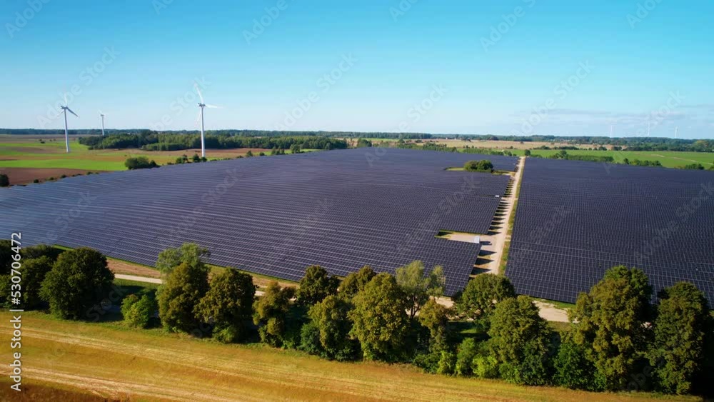 Solar Panels Farm Field Of Green Renewable Energy and Rotating Wind Turbines in background - Aerial panoramic shot
