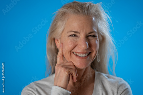 Portrait of happy smiling flirty mature woman with finger on face isolated on blue background studio. People lifestyle concept