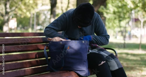 Focused homeless young woman rummaging in bag in slow motion sitting on bench in sunny park. Portrait of sad Caucasian poor houseless person searching food outdoors. Cinema 4k ProRes HQ photo