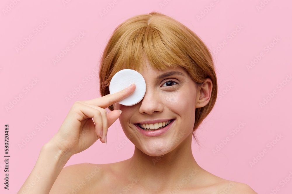 a happy, sophisticated, funny woman with blonde hair gathered back, standing on a pink background, holding a cotton pad in her hand, presses it to her eye, smiling funny a happy, sophisticated, funny woman with blonde hair gathered back, standing on a pink background, holding a cotton pad in her hand, presses it to her eye, smiling funny