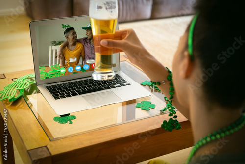 Mixed race woman holding beer having st patrick's day video call with male friends on laptop at home