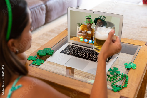 Caucasian woman holding beer having st patrick's day video call with friends on laptop at home