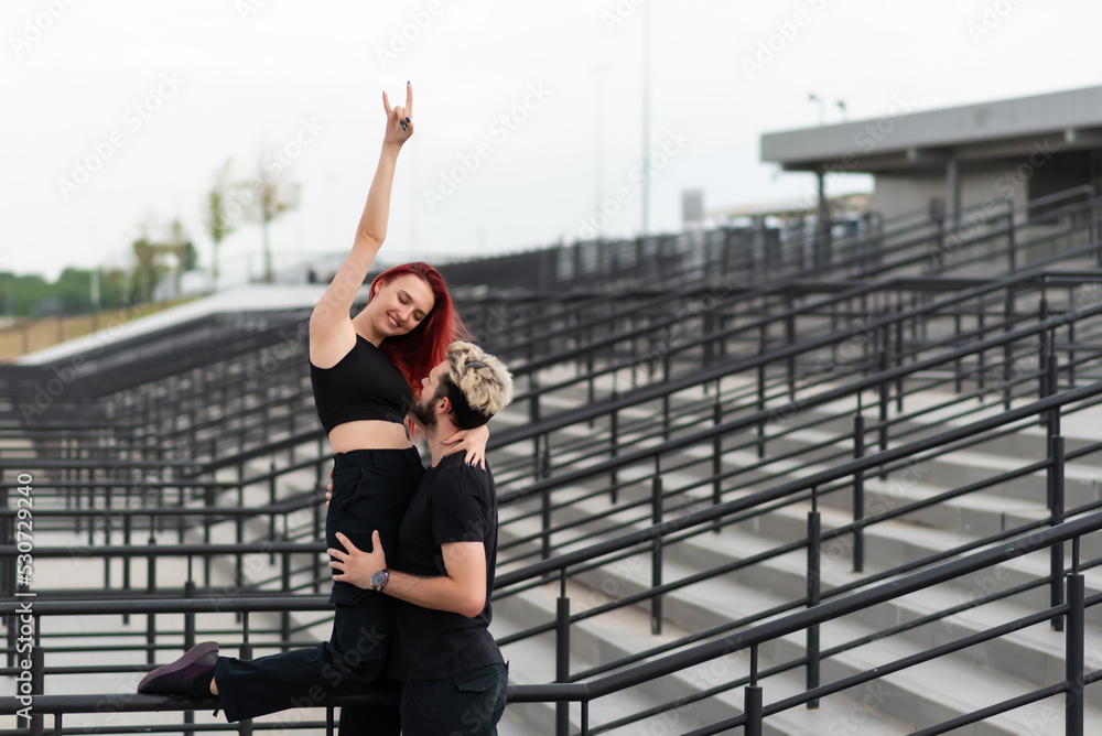 Stylish couple in love in black clothes walks in the park and hugs. Lovely couple of hipsters hugging on the background of the stairs. Emotional girl with hands up