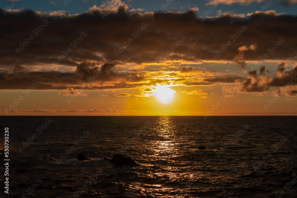 Watching the sunset at the port of Puerto de la Cruz, Tenerife, Canary Islands, Spain, Europe. View of the horizon of Atlantic Ocean. The sun is reflecting in the calm sea. Vibrant clouds in the sky