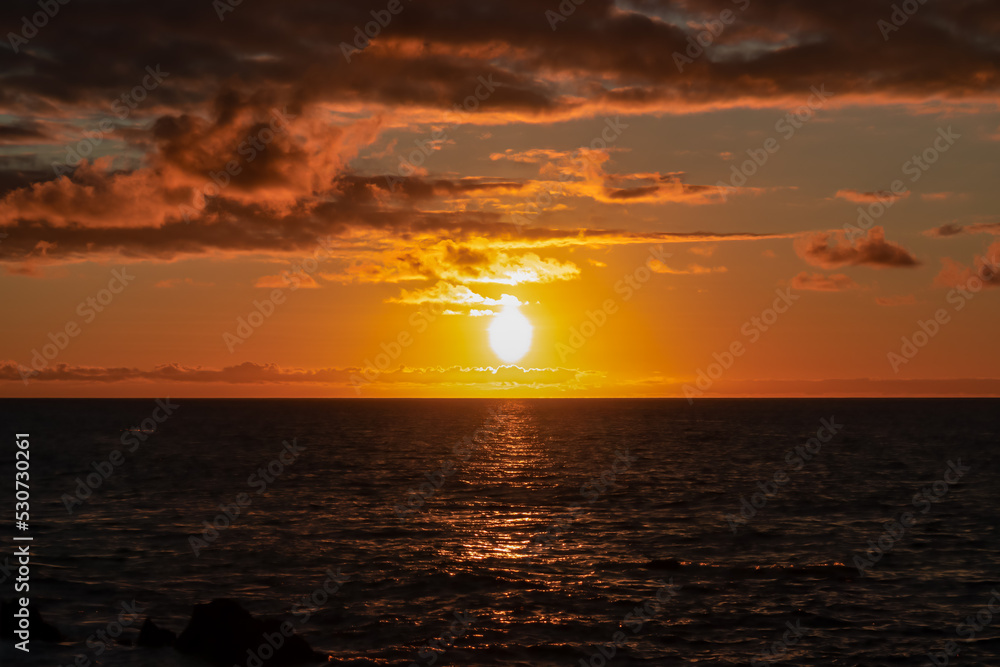 Watching the sunset at the port of Puerto de la Cruz, Tenerife, Canary Islands, Spain, Europe. View of the horizon of Atlantic Ocean. The sun is reflecting in the calm sea. Vibrant clouds in the sky