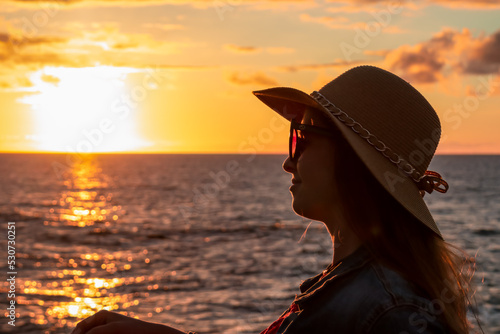 Beautiful luxury woman in summer hat and red dress sitting at port of Puerto de la Cruz during sunset, Tenerife, Canary Islands, Spain, Europe. Vacation at the sea. Reflection of sun beams in the sea