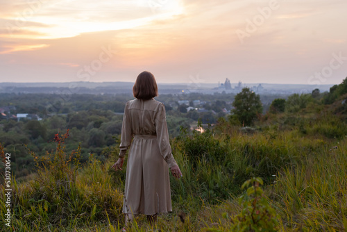 Beautiful young smiling girl in a long brown dress stands along the lawn. Happy woman walks at sunset on a hill overlooking the river. Concept of having rest in park during summer holidays or weekends