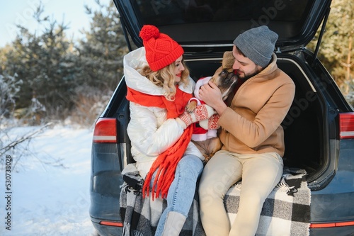 Smiling couple with dog sitting in open SUV car trunk in snowy forest. Enjoying each other in active winter holidays.