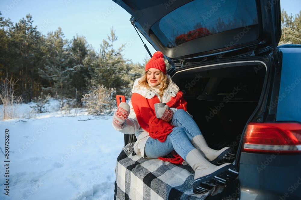 young woman sitting in car trunk drinking warm tea and takes a selfie at winter snowed day.