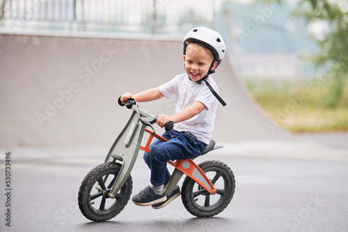 Smiling child riding balance bike. Male toddler kid in helmet learning to ride on run bicycle at skate park. © anatoliy_gleb