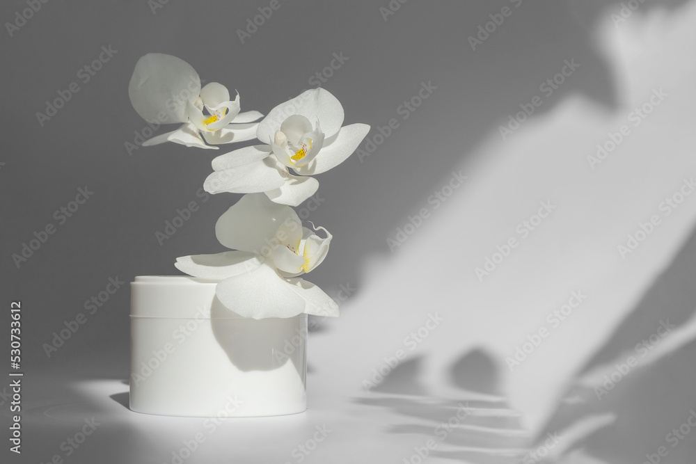 White jar of cream on light background in rays of sunlight. Packaging for cosmetic products with levitating orchid flowers. Concept of stylish appearance, mockup White jar of cream on light background in rays of sunlight. Packaging for cosmetic products with levitating orchid flowers. Concept of stylish appearance, mockup
