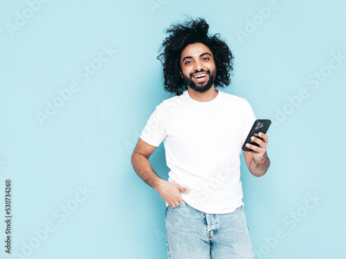 Handsome smiling hipster model.Sexy Arabian man in summer stylish clothes.Fashion male with long curly hairstyle posing near blue wall in studio.Looking at cellphone screen. Using apps