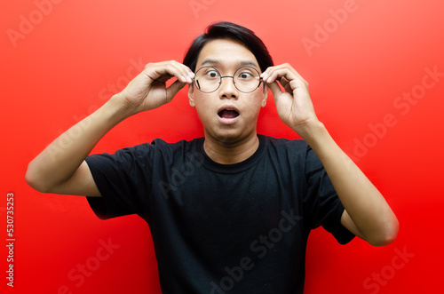Shocked face of Asian man lowering glasses in black shirt on red background.