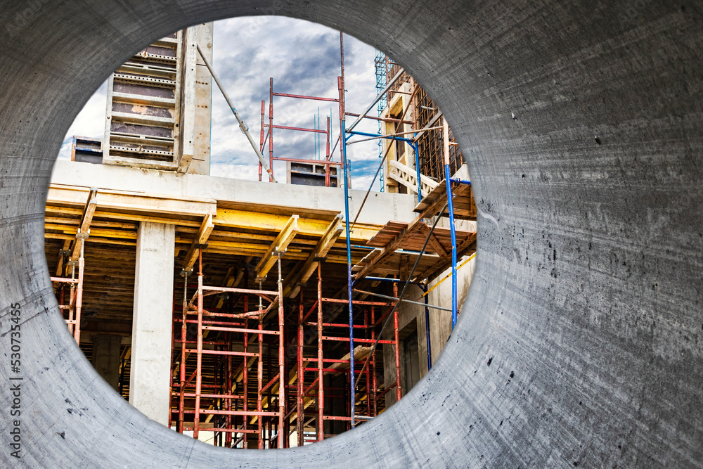 Obraz premium Construction of a new monolithic reinforced concrete house. Scaffolding on the facade of a building under construction. Working at height on a construction site. View from a large concrete pipe.