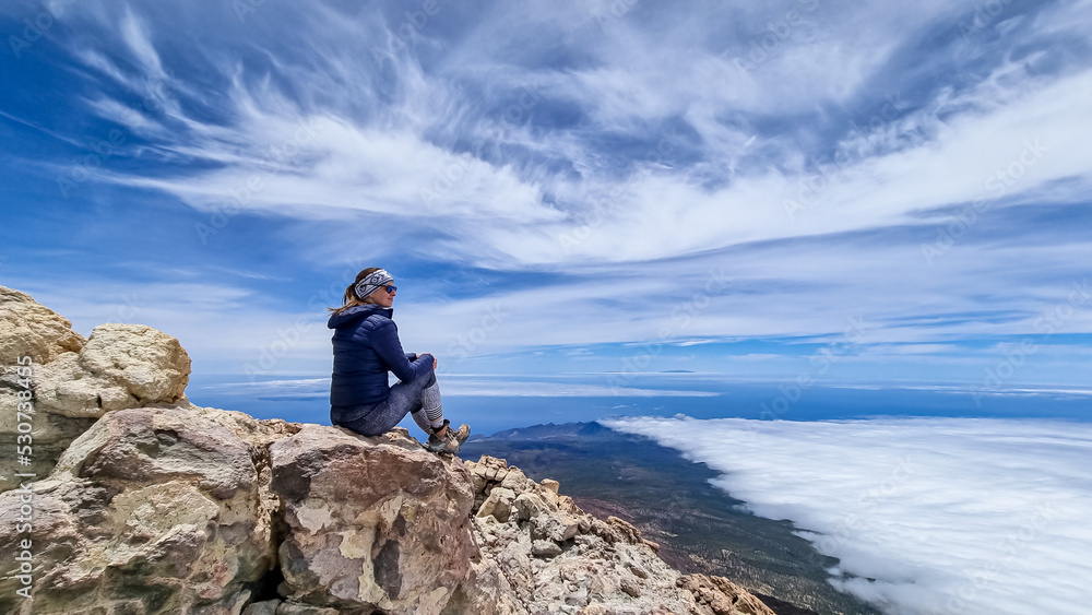 Rear view on hiking woman sitting on rock enjoying panorama from summit of volcano Pico del Teide on the island of Tenerife, Canary Islands, Spain, Europe. Island covered in cloud. Freedom concept