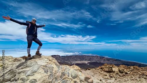 Front view of happy hiking woman in wind jacket standing on rock on summit of volcano Pico del Teide on the island of Tenerife  Canary Islands  Spain  Europe. Island covered in cloud. Freedom concept