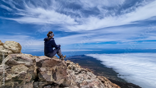 Rear view on hiking woman sitting on rock enjoying panorama from summit of volcano Pico del Teide on the island of Tenerife, Canary Islands, Spain, Europe. Island covered in cloud. Freedom concept