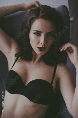 A young attractive girl with dark hair in black lingerie with evening make-up  sitting on a grey armchair  in the bedroom. beauty and fashion