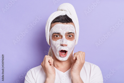 Indoor shot of shocked young adult man with mask for skin, wrapped in towel on the head isolated over violet background, looking at camera with surprised expression.