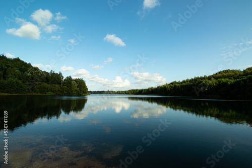 Reflection of trees in lake, blue skies with some small puffs of white clouds.