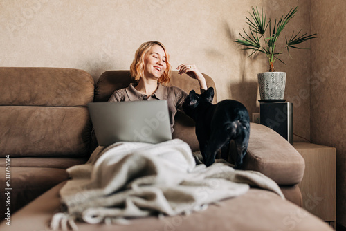 Young woman working with laptop remotely and spending day with pet in apartment interior. Front view of beautiful young woman looking at computer screen and talking to dog while sitting on the couch