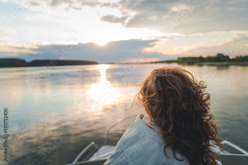 Curly young woman sitting on a boat looking at the sunset on the river, back view © olezzo