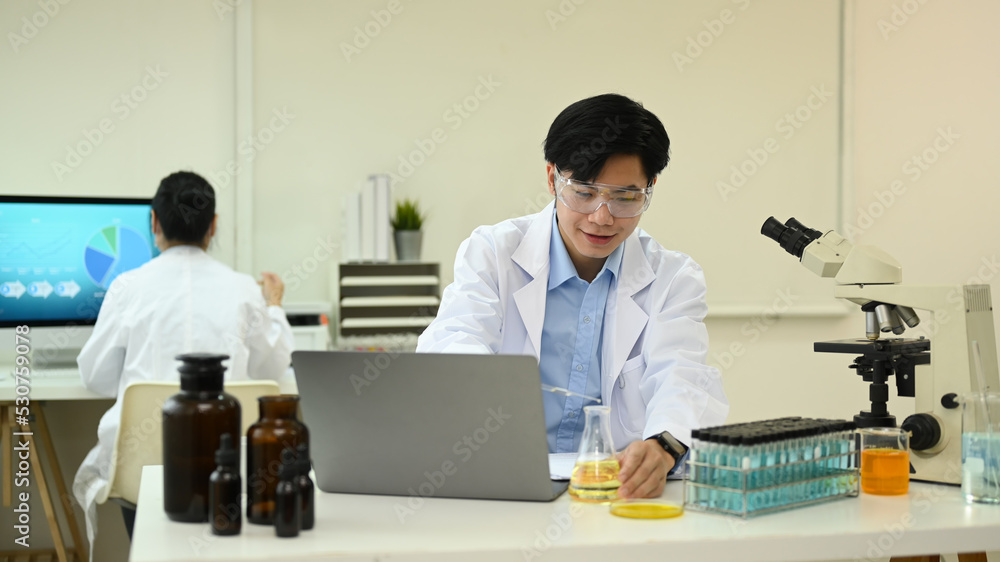 Fototapeta premium Asian man scientist in white lab coat using laptop and summarising information for scientific report