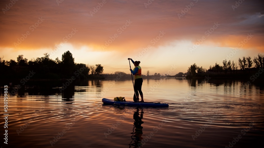 Silhouette of a young woman standing on the Sup board in the sunset lake