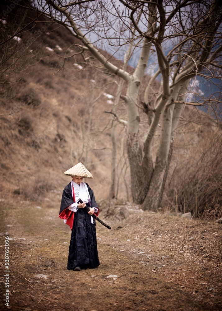 A young caucasian boy seven year old dressed as Samurai in black and white Kimono with katana standing outside in the cloudy spring mountains 