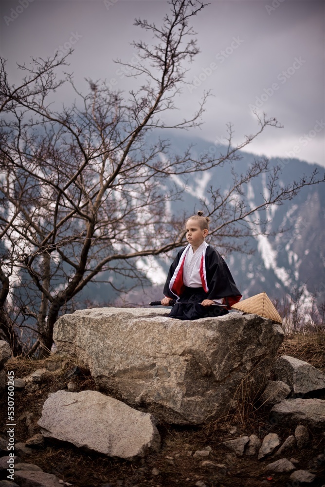 Portrait os a young caucasian boy seven year old dressed as Samurai in black and white Kimono and katana sitting outside in the mountains beside the spring tree