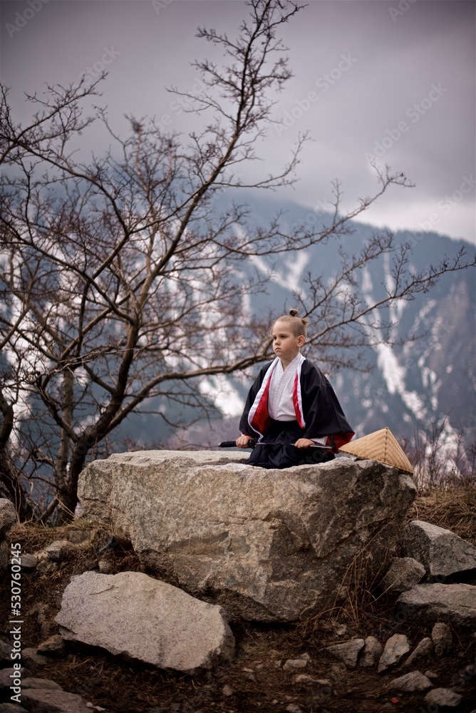 Portrait os a young caucasian boy seven year old dressed as Samurai in black and white Kimono and katana sitting outside in the mountains beside the spring tree Portrait os a young caucasian boy seven year old dressed as Samurai in black and white Kimono and katana sitting outside in the mountains beside the spring tree