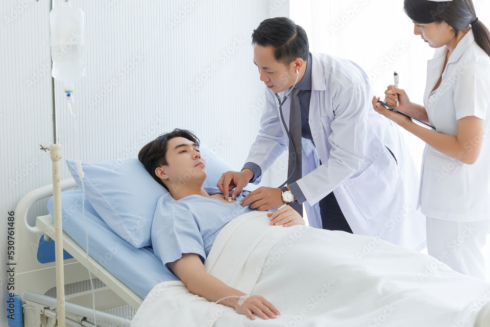Senior doctor and young male patient who lie on the bed while checking symptom, consult and explain with nurse taking note and supporting in hospital wards. Senior doctor and young male patient who lie on the bed while checking symptom, consult and explain with nurse taking note and supporting in hospital wards.
