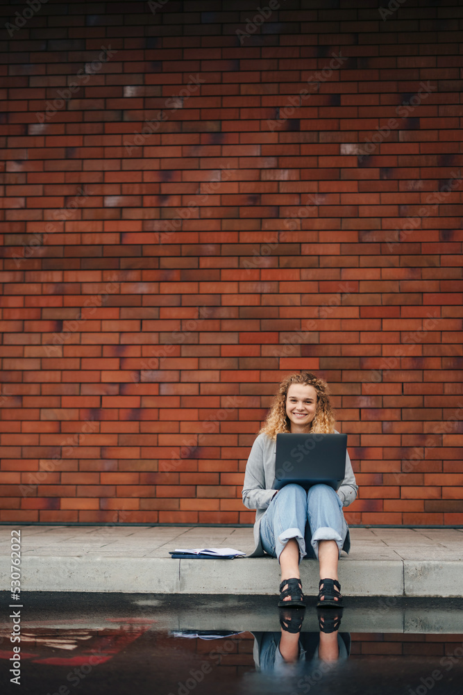 Happy young curly woman student typing on laptop sitting outdoors in city street against brick wall background with copy space. Internet technology. Modern
