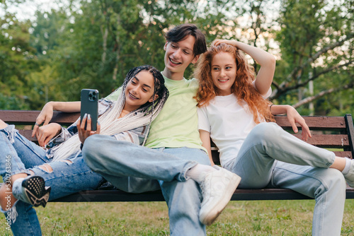 Three teenagers blogger sitting on wooden bench in the summer park using a smartphone for video communication. Modern communication. Green city. Distance © Strelciuc