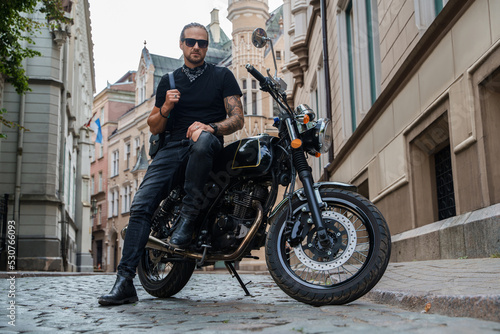 Portrait of attractive man biker with tattooes posing on his motorcyle in alleyway downtown.