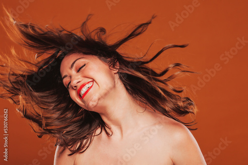 Face shot portrait of a happy smiling beautiful brunette woman in move with a smooth hair, and classic make-up. Beauty portrait at studio isolated over brown background.