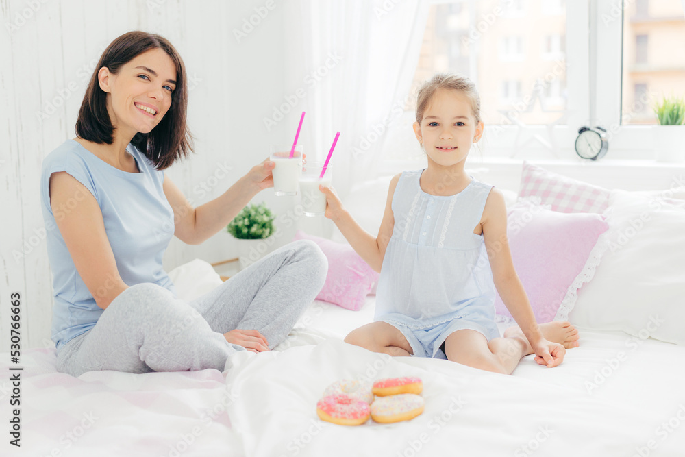 Portrait of happy young mother and her small daughter hold glasses of milk shake, have breakfast in bed, look positively at camera, enjoys weekend at home and togetherness, dressed in nightwear Portrait of happy young mother and her small daughter hold glasses of milk shake, have breakfast in bed, look positively at camera, enjoys weekend at home and togetherness, dressed in nightwear