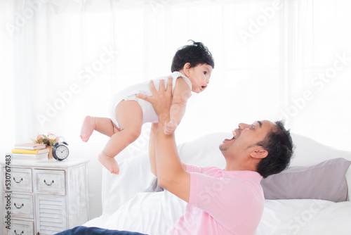 Selective focus asian father play and tease with newborn baby in bedroom. Dad holding lifting toddler on bed with fun and tender. Adorable little boy having fun with daddy togerther at home. photo