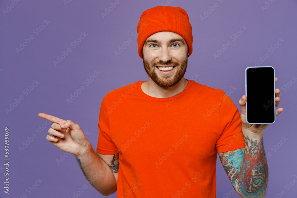 Young man wear red hat t-shirt hold in hand use mobile cell phone with blank screen workspace area point finger aside isolated on plain pastel light purple background studio People lifestyle concept. Young man wear red hat t-shirt hold in hand use mobile cell phone with blank screen workspace area point finger aside isolated on plain pastel light purple background studio People lifestyle concept.