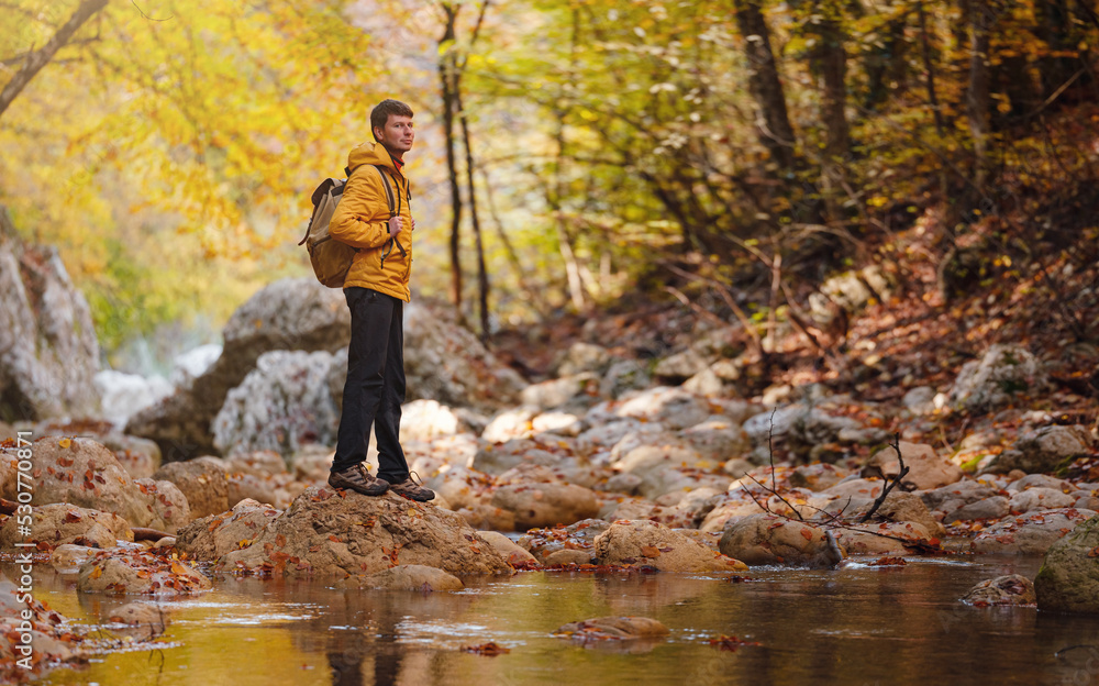 Obraz premium Travel and road trip concept at autumn. Adventure and active lifestyle in nature. Tourist hiking in forest. Caucasian man in yellow jacket walks in woods.