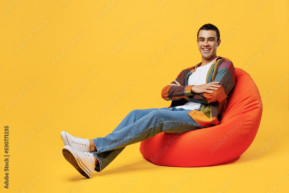 Full body young middle eastern man wear casual shirt white t-shirt sit in bag chair hold hands crossed folded look camera isolated on plain yellow background studio portrait People lifestyle concept. Full body young middle eastern man wear casual shirt white t-shirt sit in bag chair hold hands crossed folded look camera isolated on plain yellow background studio portrait People lifestyle concept.
