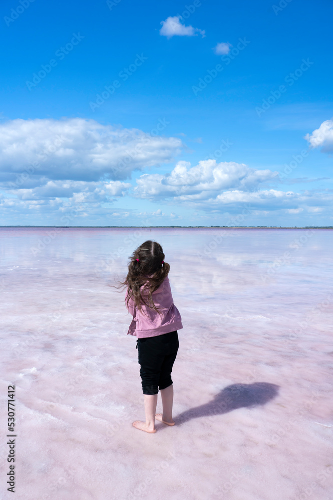 Bursol pink lake. A girl with ponytails stands on the shore of a salt lake and looks into the distance. Clouds are reflected in pink water. Concept: freedom, looking into the bright future, lightness